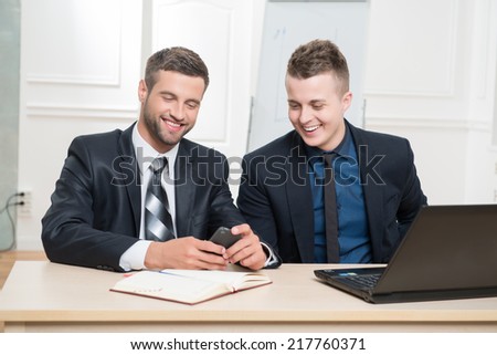 Waist-up portrait of two handsome businessmen in suits sitting at the table in office interior, one holding in his hands a mobile phone and both lightly laughing while looking on the screen of phone
