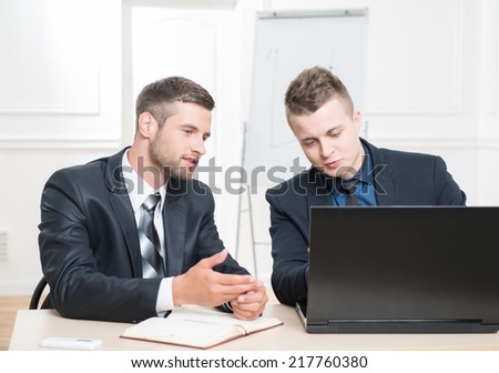 Waist-up portrait of two handsome businessmen in suits sitting at the table with laptop in office interior seriously looking on the screen of laptop while discussing a new project
