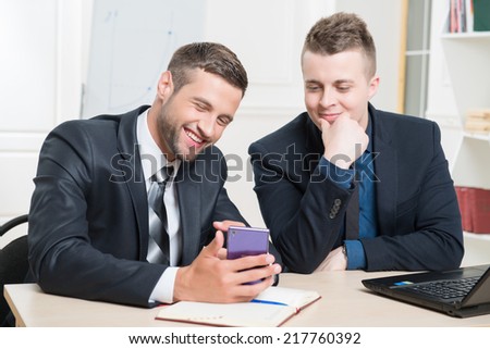 Waist-up portrait of two handsome businessmen in suits sitting at the table in office interior, one holding in his hands a mobile phone and both lightly laughing while looking on the screen of phone