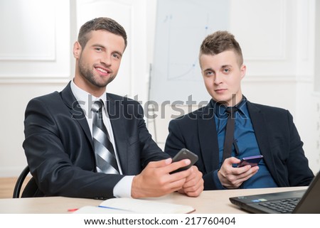 Waist-up portrait of two handsome businessmen in suits sitting at the table and holding in their hands mobile phones and looking at the camera with a nice smile in office interior