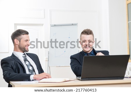 Waist-up portrait of two handsome businessmen in suits sitting at the table with laptop in office interior, laughing and looking on the screen of laptop while discussing a new project