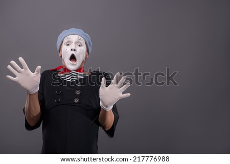 Waist-up Portrait of young male mime confused breeding his hands showing that he is scared and frighten looking at you with wide opened mouth and eyes isolated on grey background with copy place