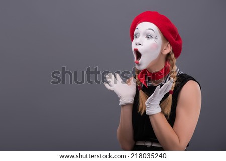 Waist-up portrait of young mime girl with red hat and red scarf looking aside with pleasant surprise, showing that she is happy to see something, isolated on grey background with copy place