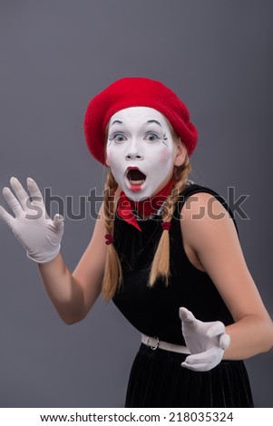 Waist-up portrait of young mime girl with red hat and red scarf looking at the camera with sudden fright and waving her hands isolated on grey background with copy place