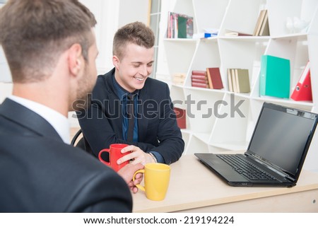 Close-up portrait of two handsome businessmen in suits having a coffee-break in office sitting at the table, with selective focus on one smiling businessman and another sitting back to the camera