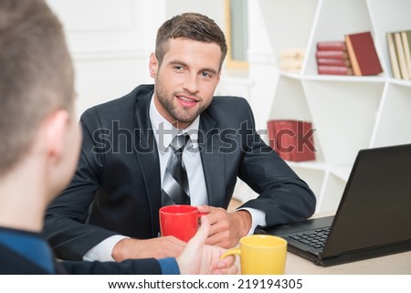 Close-up portrait of two handsome businessmen in suits having a coffee-break in office sitting at the table, with selective focus on one smiling businessman and another sitting back to the camera