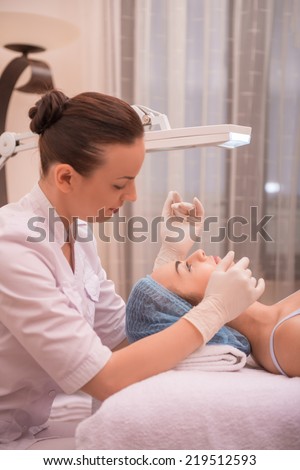 Side-view portrait of cosmetologist and a young woman with fresh and clean skin closing her eyes and lying on a table getting a professional cosmetology procedures in healthy beauty spa salon