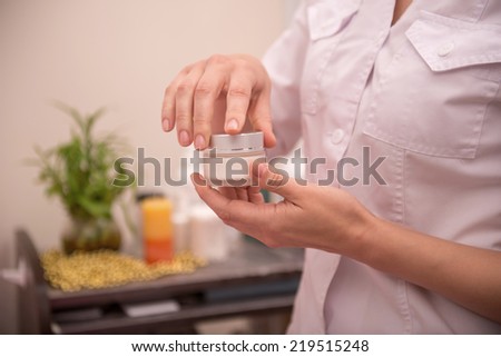 Close-up picture of hands of a young cosmetologist in healthy beauty spa salon interior holding in her hands and opening a bottle with facial cream