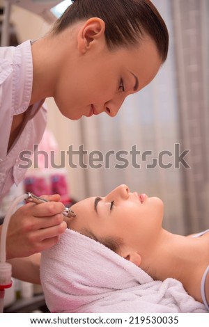 Side-view portrait of a cosmetologist and a young woman with a towel on her head lying on a table with closed eyes getting a laser skin treatment in healthy beauty spa salon