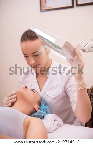 Side-view portrait of cosmetologist and a young woman with fresh and clean skin closing her eyes and lying on a table getting a professional cosmetology procedures in healthy beauty spa salon