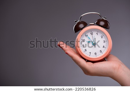 Waist-up portrait of pretty girl with surprised face holding an alarm clock in her hand and looking at it with opened mouth showing at a clock, with copy place isolated on grey background