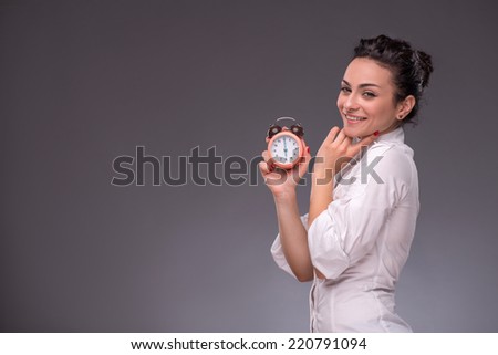 Waist-up portrait of beautiful girl thinking about something  and smiling while holding an alarm clock and looking at you, with copy place isolated on grey background concept of time management