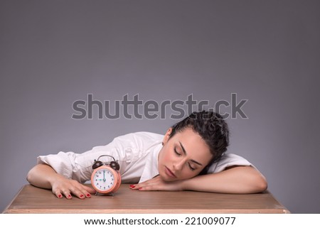 Waist-up portrait of young girl sitting at the table with small alarm clock and holding her hands on the table confident looking at the camera, isolated on grey background with copy place