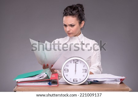 Waist-up portrait of girl sitting at the table in office with a pile of documents and the clock and concentrated reading some papers isolated on grey background with copy place