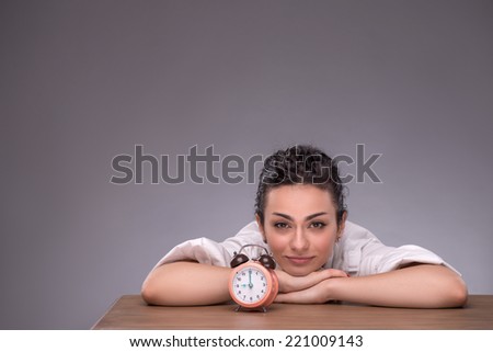 Waist-up portrait of young girl sitting at the table with small alarm clock and holding her head on her hands on the table, calmly looking at the camera isolated on grey background with copy place