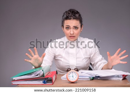 Waist-up portrait of girl sitting at the table with documents and an alarm clock, breeding her hands because she has no time, confused looking at you, isolated on grey background with copy place