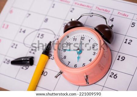 Close-up top-view photo of calendar with a datum circled with a black marker lying on it, with selective focus on an alarm clock, concept of time management at work