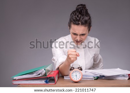 Waist-up portrait of girl sitting at the table in office with a pile of documents and an alarm clock wanting to crash it because she has no time isolated on grey background with copy place