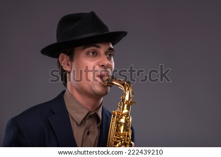 Waist-up portrait of a young handsome Caucasian jazz man in a suit with a black hat looking at the camera with astonishment holding a trumpet in his hand isolated on grey background with copy place