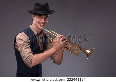 Waist-up portrait of a young handsome Caucasian jazz man in a suit with a black hat looking at the camera holding a trumpet in his hand and playing on it, isolated on grey background with copy place