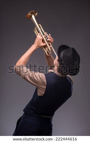 Waist-up portrait of a jazz man in a suit with a black hat hiding his face and standing back to the camera while playing a trumpet isolated on grey background with copy place, concept of jazz music
