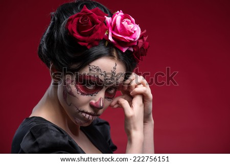 Close-up portrait of girl with Calaveras makeup and a red flower in her black hair looking at you under a forehead and holding her hands near her face isolated on red background with copy place