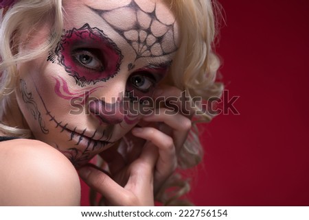 Close-up portrait of young blond girl with sad face with Calaveras makeup upset looking at the camera while holding her hands near face isolated on red background with copy place