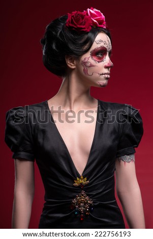 Waist-up portrait of young girl in black dress with Calaveras makeup and a red flower in her black hair looking aside isolated on red background