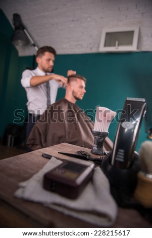 hairdressing equipment in  forefront  and  hairdresser  cuts   hair  with scissors on crown of handsome satisfied  client in background  in  professional  hairdressing salon