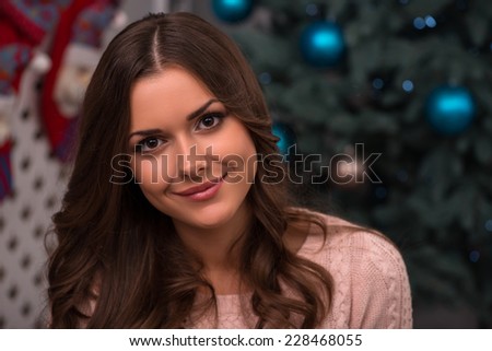 Beautiful attractive brown haired girl sitting near red Christmas socks for presents and fir tree with present in blue gift wrapping on white pile carpet dressed in beige knitted jacket