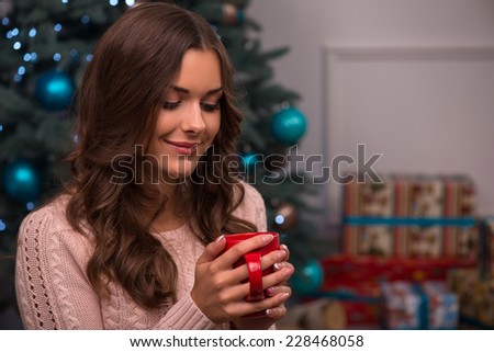 Beautiful attractive brown haired girl drinking cup of coffee or tea or milk sitting near fir tree and heap of  presents  dressed in beige knitted jacket selective focus  looking at cup