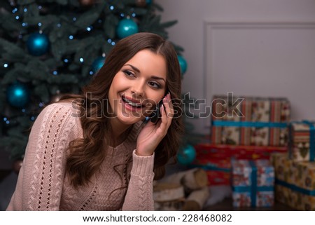 Beautiful attractive brown haired girl sitting fir tree and heap of  presents  in different  gift wrapping on white pile carpet speaking  on  phone looking at camera