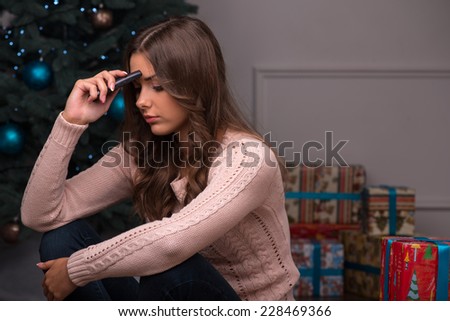 Beautiful attractive sad  disappointed brown haired girl sitting  with phone near  fir tree and heap of  presents  in different  gift wrapping on white pile carpet