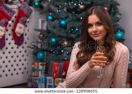 Beautiful attractive brown haired girl sitting with two glasses of champagne near red Christmas socks for presents and fir tree on white pile carpet dressed in beige knitted jacket