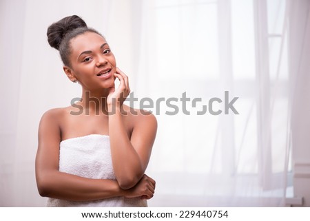 Portrait of beautiful  dark skinned girl with in white towel  in bun smiling  and touching her face  looking at camera isolated on white background  with copy place