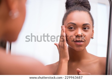 Beautiful  dark skinned girl in white towel bringing face cream looking at mirror  isolated on white background