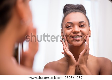 Beautiful  dark skinned girl in white towel bringing face cream looking at mirror admiring  herself  touching cheeks smiling  isolated on white background