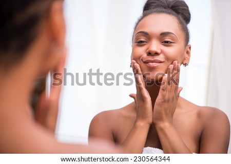 Beautiful dark skinned girl in white towel bringing face cream looking at mirror admiring  herself  touching cheeks smiling  isolated on white background