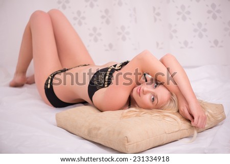 Sexy beautiful blond girl in black underclothes with blue eyes lying on her back   on white spread on beige pillow looking at camera  isolated on white background