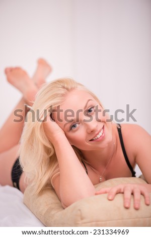 Sexy beautiful blond girl in black underclothes with blue eyes smiling  lying on stomach   on white spread on beige pillow looking camera  isolated on white background