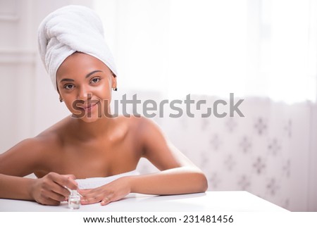 Portrait of beautiful  dark skinned girl in white towel on head and body painting her nails with transparent enamel looking at camera sitting at white table   with copy place