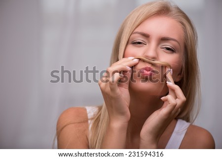 Portrait of beautiful  funny nice happy blond girl with green  eyes  and straight  hair in white  T shirt  looking at camera holding hair as moustache