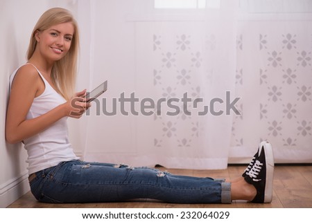 Beautiful happy  blond girl in white  T shirt  and jeans and trainers smiling looking at camera sitting on floor with  tablet computer with copy place full length