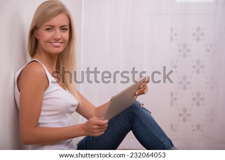 Beautiful happy  blond girl in white  T shirt  and jeans and trainers smiling looking at camera sitting on floor with  tablet computer with copy place waist up
