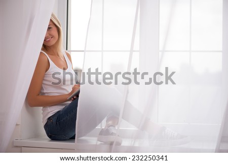 Beautiful happy  blond girl in white  T shirt  and jeans smiling  sitting on windowsill with  tablet computer looking at camera with copy place waist up