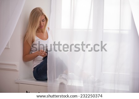 Beautiful happy  blond girl in white  T shirt  and jeans smiling  sitting on windowsill looking at  tablet computer with copy place  waist up