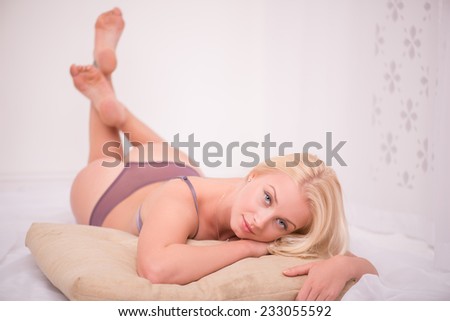 Sexy beautiful blond girl in violet  underclothes with blue eyes lying on her back   on beige pillow looking at camera touching her face  isolated on white background