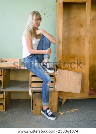 Beautiful stylish daring hussy  blond girl with straight hair in white  T shirt and jeans  confidently  proudly looking at camera  sitting on  old table  full length