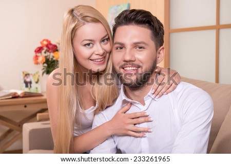 Portrait of happy couple in love of handsome man and attractive woman embracing him sitting on sofa looking at camera waist up