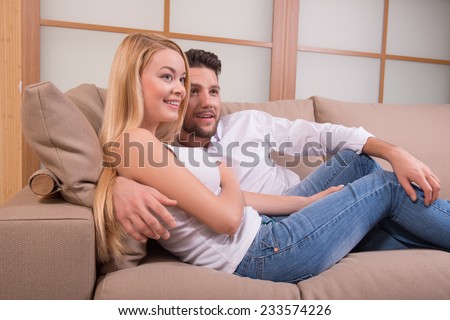 Portrait of happy couple in love of handsome man closing eyes  and attractive woman sitting on sofa embracing smiling looking aside waist up